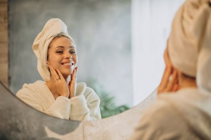 Mulher sorrindo com roupão e toalha na cabeça se olhando no espelho